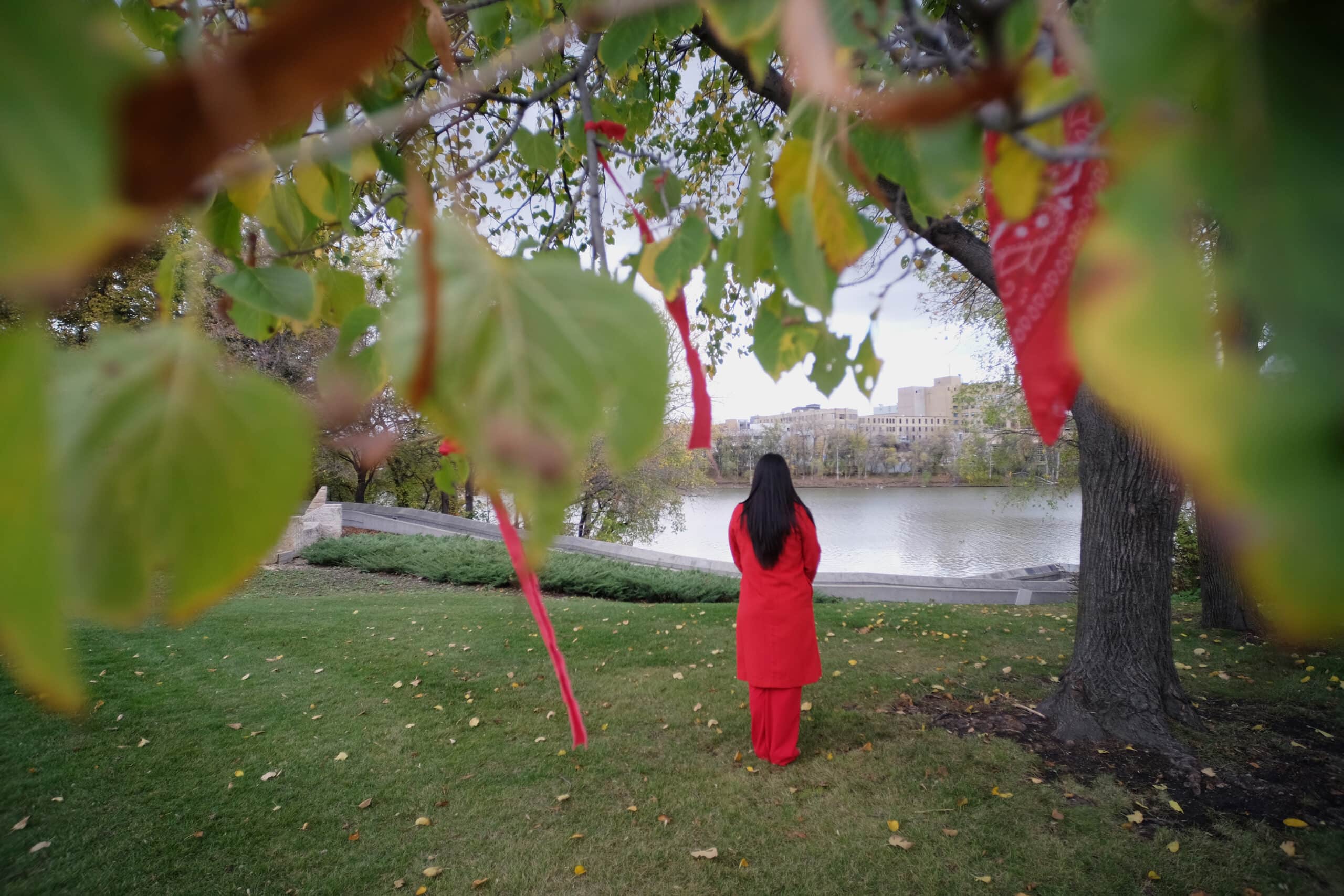 Une femme aux longs cheveux foncés, vêtue de rouge, regarde vers la rivière Rouge à l'arrière-plan de la photo, tandis que des rubans rouges commémoratifs attachés aux branches des arbres sont au premier plan.